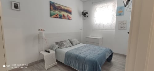 Luminoso dormitorio moderno en Apartamentos Huelintown, Málaga, con ropa de cama azul, cabecero blanco y ventilador