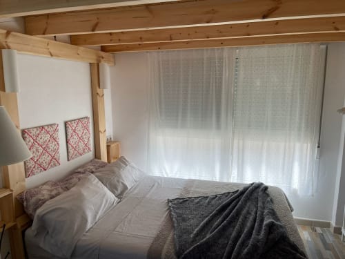 Dormitorio luminoso en Apartamentos Huelintown, Málaga, con vigas de madera, cabecero rojo estampado y cortinas translúcidas