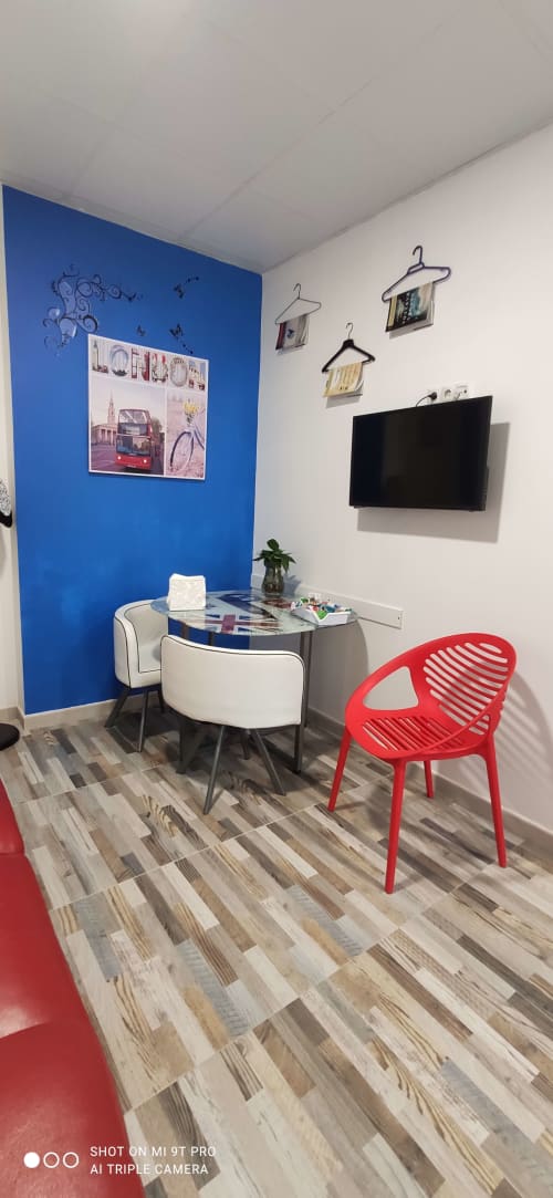 Estudio moderno en Apartamentos Huelintown en Málaga con pared de acento azul, mesa de cristal redonda, silla roja y lámpara de pared