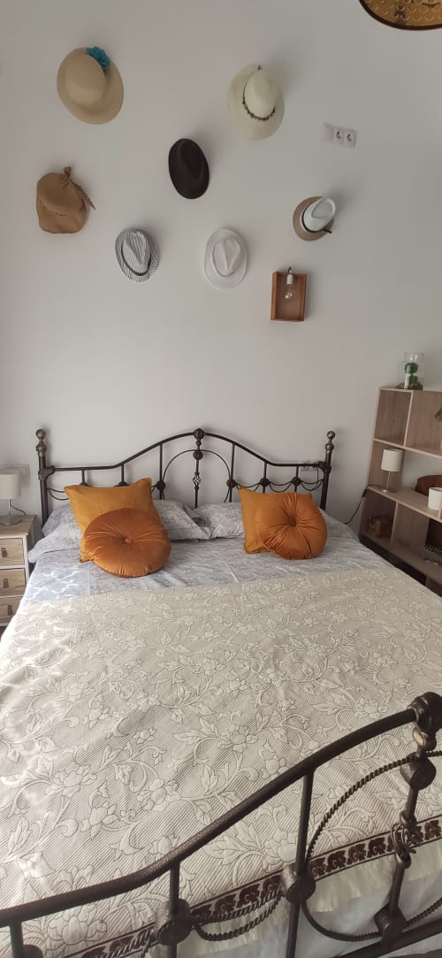 Dormitorio en Apartamentos Huelintown, Málaga con cama de hierro, almohadas naranjas y decoración de sombreros en la pared