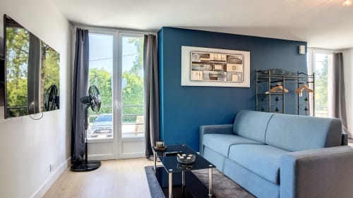 Salon moderne à la Résidence L'Hermitière avec mur d'accent bleu, canapé gris clair, TV et vue sur le jardin