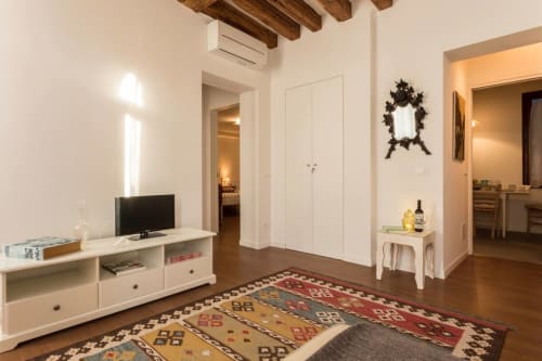 Modern apartment living room in Venice with wooden beams, white cabinetry, TV, decorative mirror, and patterned rug