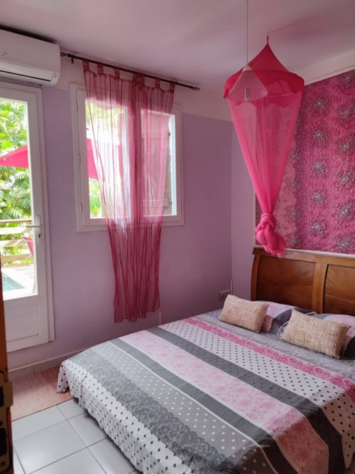 Chambre rose avec lit en bois, rideaux voilage rouges, baldaquin, papier peint floral et accès au balcon à la Résidence Les 2 Moulins,