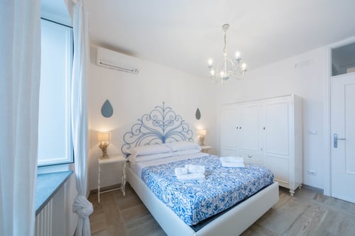 Camera da letto luminosa e moderna a Villa Costanza sulla Costiera Amalfitana con testata ornamentale blu, arredi bianchi e finestra con vista mare