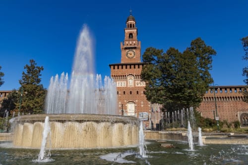 Fontana del Castello Sforzesco a Milano con torre dell'orologio e alberi verdi sotto cielo azzurro