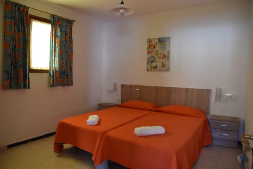 Habitación luminosa en Pension Eva, Puerto de Mogán, con dos camas, ropa de cama naranja, cortinas estampadas y decoración sencilla