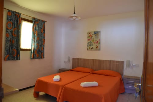 Luminosa habitación hostal en Pension Eva con camas gemelas, ropa de cama naranja, cortinas estampadas y cabecero de madera en Puerto de Mogán
