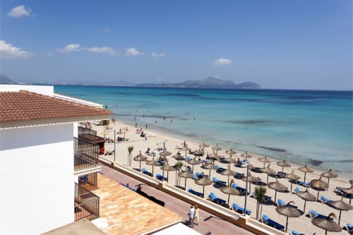 Hotel Sa Roqueta beachfront view with straw umbrellas, blue loungers, and turquoise Mediterranean sea in Can Picafort