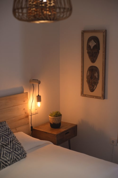 Cozy bedroom corner at La Casa De La Abuela Regina with wooden nightstand, pendant lamp, potted plant, and tribal mask wall