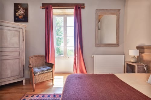 La Vieille Dame cottage bedroom with purple bedding, wicker chair, mountain views through tall windows with red curtains, and