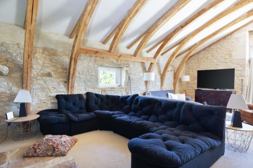 Lounge loft contemporain avec canapé sectionnel bleu marine, poutres apparentes, murs en pierre et écran plat aux Lumieres Du Causse