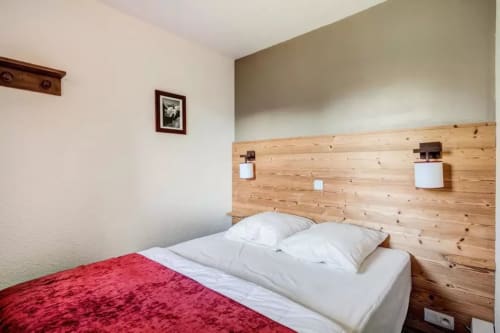 Chambre moderne au CMD Méribel Le Pralin avec tête de lit en bois, oreillers blancs, couverture rouge et éclairage intégré