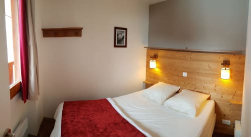 Chambre moderne à CMD Méribel Le Pralin avec tête de lit en bois, literie blanche, plaid rouge, éclairage chaleureux et vues sur la montagne