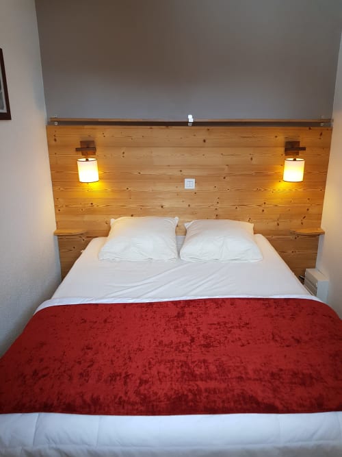 Chambre moderne au CMD Méribel Le Pralin avec tête de lit en bois, oreillers blancs, jeté rouge en velours et appliques murales