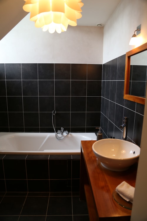 Modern bathroom at Le Grand Duc in Valenciennes with black tile walls, white bathtub, and wooden vessel sink under warm