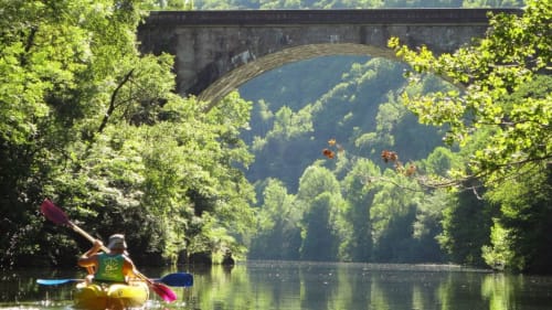 Kayakiste pagayant sur une rivière paisible sous un pont en pierre, entouré d'arbres verdoyants près des Lumières du Causse
