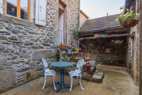 Cour de pierre charmante au Refuge des Alpes Mancelles avec chaises de bistrot blanches, table bleue et plantes fleuries