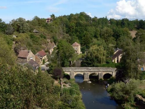 Medieval stone bridge over river with hillside cottages and lush greenery in Fresnay-sur-Sarthe
