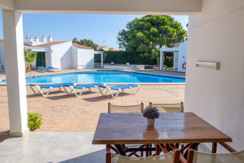 Luminosa zona de piscina en Las Golondrinas con tumbonas, mesa de comedor y apartamentos blancos con vistas al jardín mediterráneo