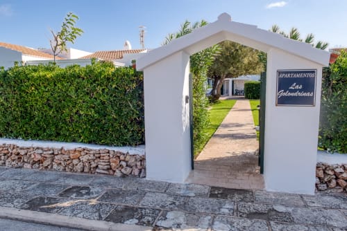 Puerta de entrada a apartamentos Las Golondrinas con pórtico blanco, pasillo ajardinado y setos en Cap d'Artrutx