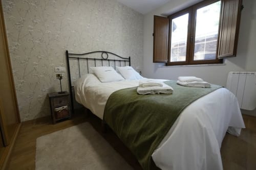 Luminosa habitación en Vivienda Balbino con cama de hierro forjado, ropa blanca, manta verde, papel tapiz estampado y vistas a la montaña