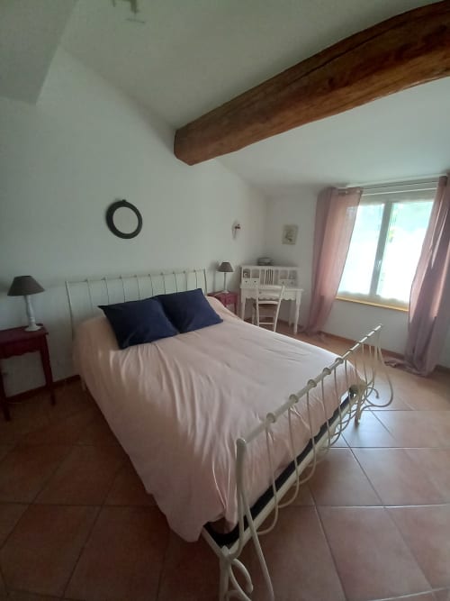 Chambre lumineuse au gîte Sylvie Cambon à Montséret avec lit en fer blanc, coussins bleu marine, plafond poutre bois et vue jardin