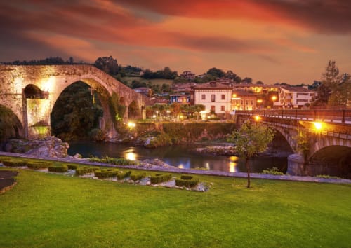 Puente de piedra histórico y edificios antiguos reflejados en el río al atardecer, La Venta de Miyares en Piloña, España