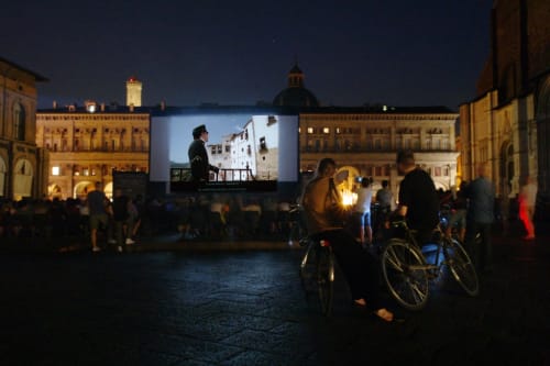 Outdoor movie screening in Bologna's historic piazza with cyclists and locals watching, illuminated Renaissance buildings at