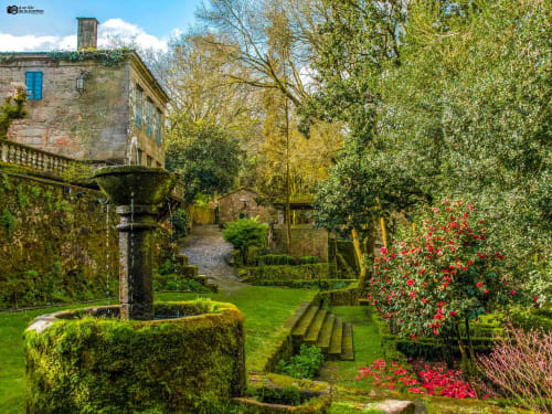 Camino de jardín exuberante en Cabañas Compostela, Santiago de Compostela, con edificios de piedra, muros cubiertos de hiedra y flores rojas