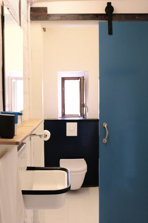 Baño moderno con inodoro y lavabo negros, paredes color crema y puerta azul en Casa rural MAÑARINEGI de Gipuzkoa