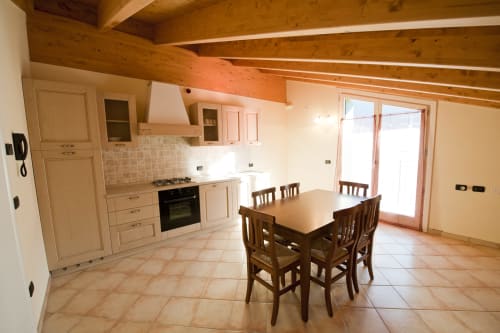 Cucina-soggiorno moderna con travi in legno, ante color panna, tavolo da pranzo e porte vetrate che si aprono sul balcone della Residenza