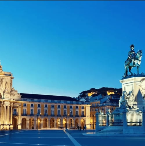 Praça histórica de Leiria ao anoitecer com edifícios coloniais amarelos, estátua equestre e castelo iluminado no topo da colina