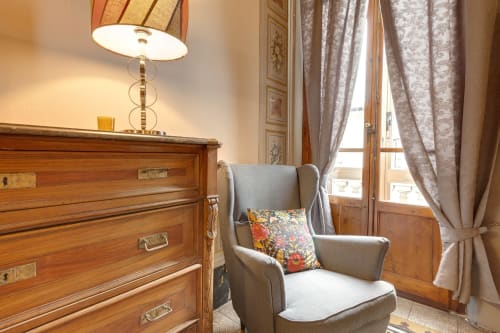 Elegante angolo camera da letto presso Sleep tight Florence con comò in legno, poltrona wingback grigia, cuscino decorativo e