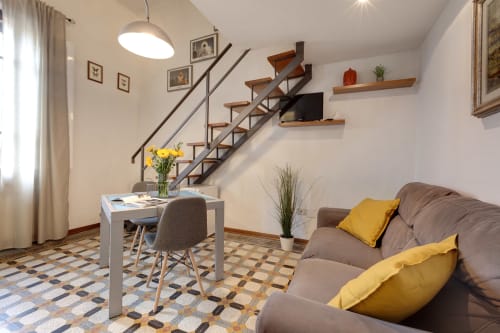 Loft a Firenze con divano grigio, cuscini gialli, tavolo da pranzo e scala industriale con ripiani in legno