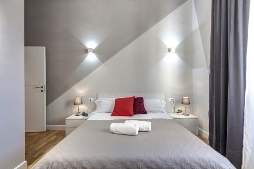 Camera da letto moderna in mansarda presso Sleep tight Florence con letto bianco, cuscini rossi, soffitto inclinato, applique da parete e rivestimenti in legno