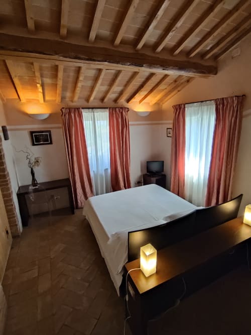 Camera rustica a La Locanda del Vino Nobile a Montepulciano con travi in legno a vista, letto matrimoniale, tende rosse e atmosfera calda