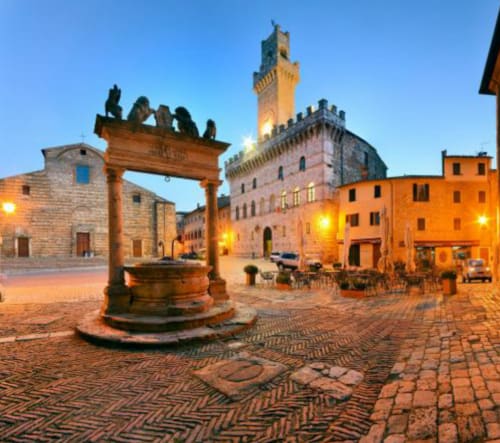La storica piazza di Montepulciano al tramonto con torre rinascimentale, pozzo e edifici in pietra illuminati da calde luci