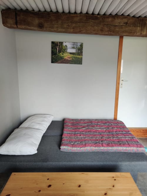 Chambre de l'appartement La Ferme De Werpin avec lit, oreiller blanc, couverture rouge, poutres en bois et tableaux paysagers