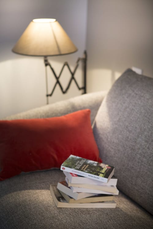 Coin de lecture douillet à La Maison De Siloe avec livres empilés, coussin rouge, lampe et fauteuil gris rembourré.