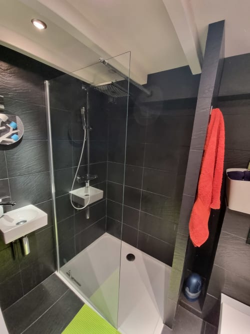 Salle de bain moderne aux murs en carrelage noir, pomme de douche pluie, paroi vitrée, serviette rouge et sanitaires blancs à Locmaria