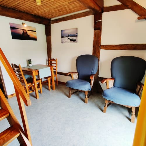 Cozy sitting area with wooden beams, two blue armchairs, dining table, and landscape photos at La Ferme De Werpin apartment