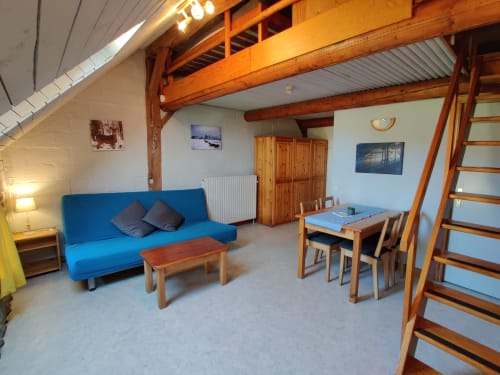 Lumineux séjour avec canapé bleu, poutres apparentes, table à manger et escalier de mezzanine à La Ferme De Werpin
