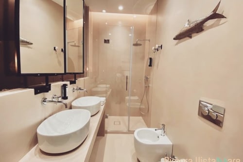 Moderno bagno di Dimora Vistamare con doppio lavabo, doccia in vetro e decorative opere d'arte con pesci su pareti color crema