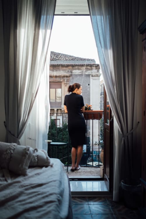 Donna in abito nero sul balcone con vista sul panorama di Catania, camera dell'hotel Sicilianinsta Rooms con tende trasparenti