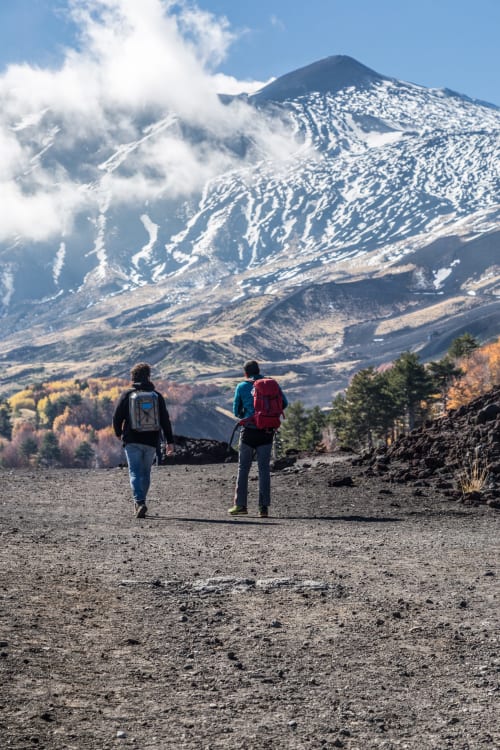 Due escursionisti con zaini su un terreno vulcanico di fronte all'Etna innevato, Sicilia