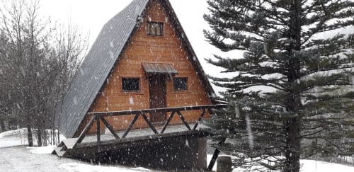 Chalet rustique en brique rouge aux toits enneigés, entouré de sapins lors d'une chute de neige à Jausiers, France.