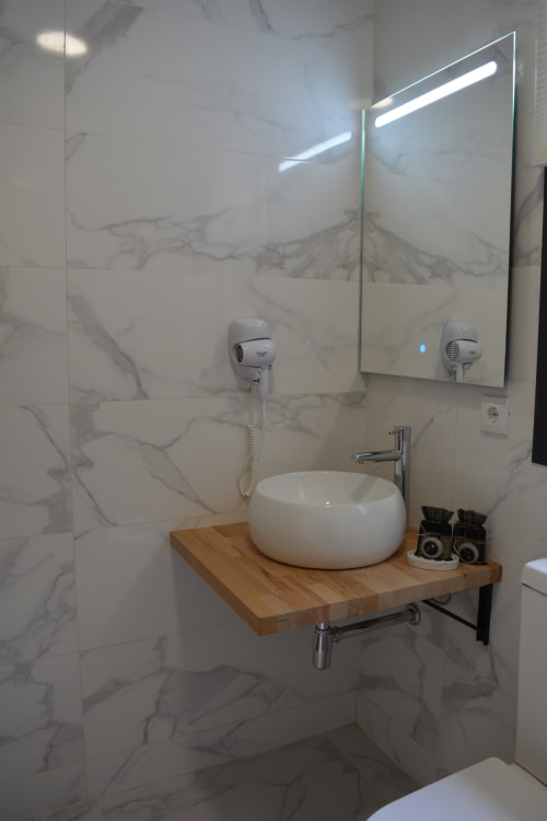 Moderno tocador de baño con lavabo redondo blanco sobre encimera de madera, espejo iluminado y azulejos de mármol en Cabañas Compostela