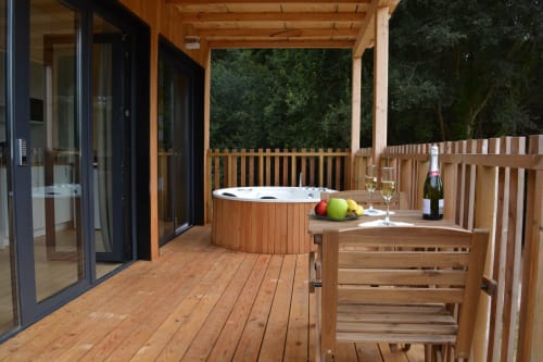 Terraza de madera con jacuzzi, mesa de comedor, botella de vino y frutas con vista al bosque verde en Cabañas Compostela