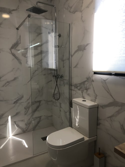 Baño moderno en apartamento Cabañas Compostela con paredes efecto mármol, ducha tipo lluvia, cristalería y acabados blancos