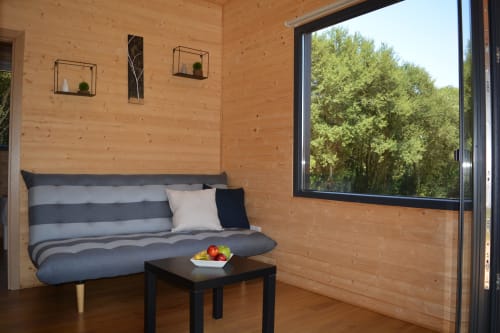 Acogedor dormitorio de cabaña de madera en Cabañas Compostela con cama rayada, mesita negra, plantas en maceta y vista al bosque